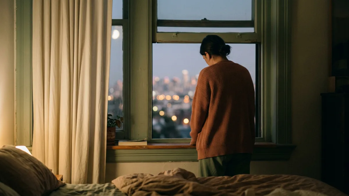 Person standing at a bedroom window above a city at dusk, representing insomnia and sleep disorder care at Mind Link